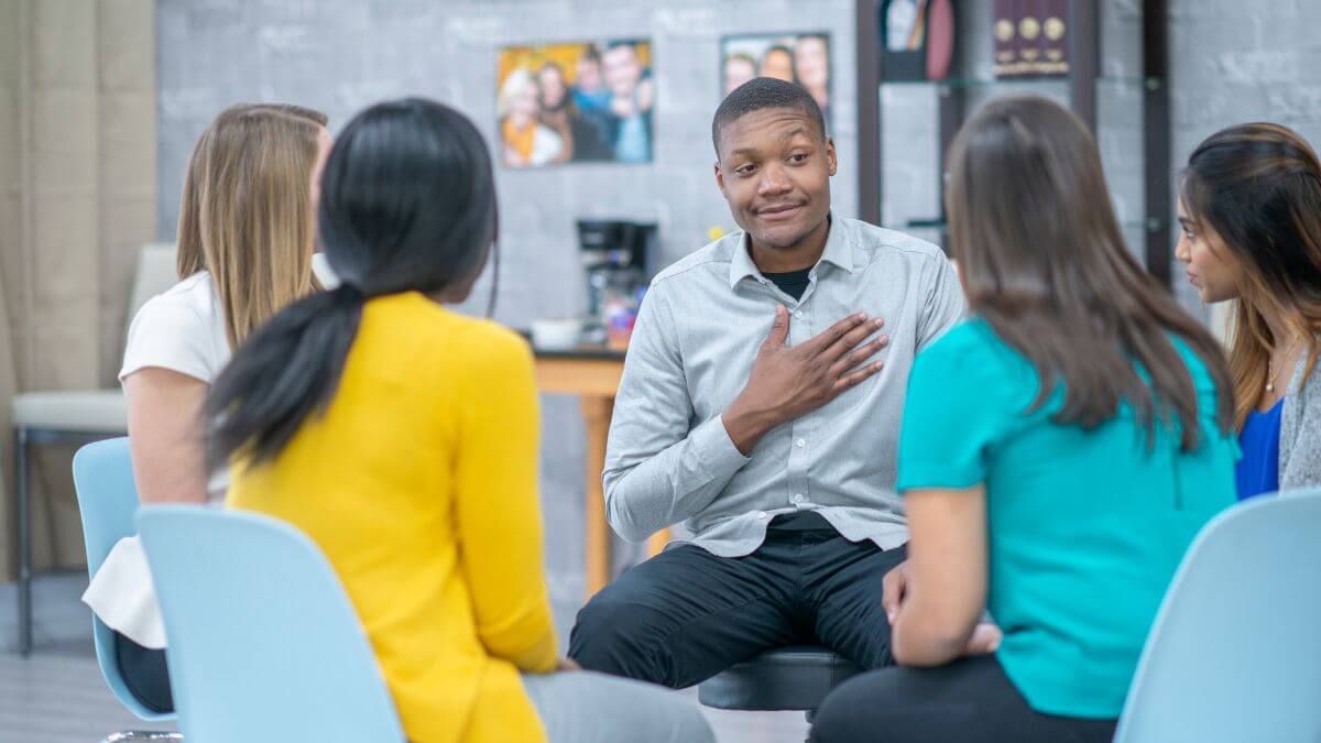 A group of five people is sitting in a circle, engaged in a discussion. A man in a gray shirt is speaking while the others listen attentively. The setting appears to be an informal meeting or support group.