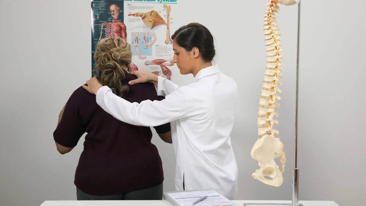 A healthcare professional is examining the back of a patient, with an anatomical poster in the background and a model of a spine on display.