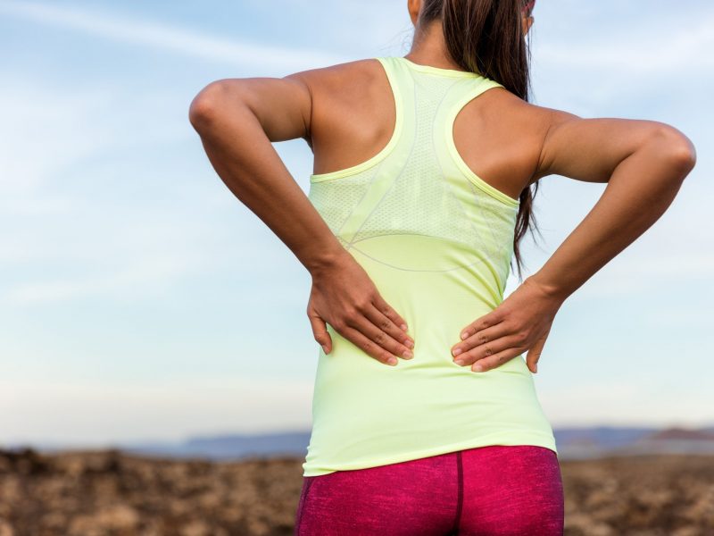 Trail running runner with painful lower back pain injury or strained muscle near the spine. Female athlete from behind on outdoor run pressing body with hands for muscles cramping soreness.